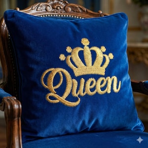 Könnte beinhalten: Ein königsblaues Samtkissen mit goldgesticktem Text und einem Kronen-Design. Das Wort "Queen" ist in einer Schreibschrift unter der Krone gestickt. Das Kissen ist quadratisch und liegt auf einem Holzstuhl.