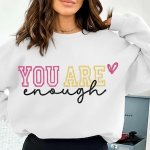 Könnte beinhalten: Weißes Sweatshirt mit dem Text "YOU ARE enough" in Rosa, Gelb und schwarzer Schrift. Ein rosa Herz befindet sich neben dem Wort "ARE". Das Sweatshirt ist ein lässiger, bequemer Stil.