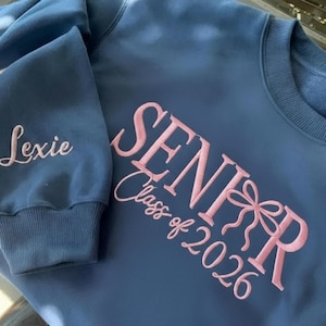 May include: A blue sweatshirt with the word "SENIOR" and "Class of 2026" embroidered in pink script. The name "Lexie" is embroidered on the sleeve in a matching pink thread. The sweatshirt has a crew neck and ribbed cuffs.