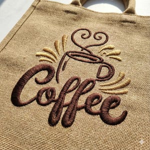 May include: A beige burlap tote bag featuring a brown embroidered coffee cup design and the word "Coffee" in a cursive font. The design includes steam and coffee cup details in brown and tan.
