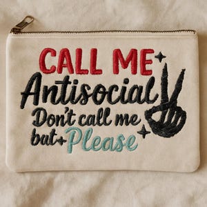 May include: Cream-colored zippered pouch with embroidered text reading "CALL ME Antisocial Don't call me but Please" in red, black, and teal. A black peace sign is embroidered on the right side. The zipper is gold.