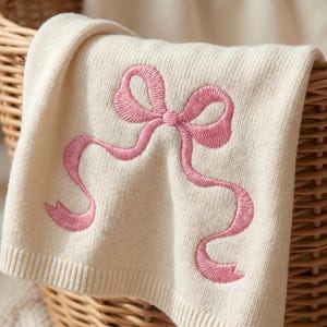 May include: Cream-colored knitted blanket with a large pink embroidered bow design. The blanket is folded over a wicker basket, showcasing the detailed stitching of the bow. The soft texture and color palette suggest a baby blanket or nursery item.