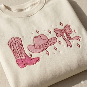 May include: Cream-coloured sweatshirt with embroidered pink cowboy-themed designs. The design includes a cowboy boot, a cowboy hat with star details, and a bow. Pink diamond and dot accents surround the images.