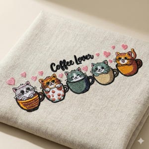 May include: Embroidered linen towel featuring five cartoon cats in coffee cups with pink hearts. The text "Coffee Lover" is embroidered above the cats. The cats are in various colors, including orange, gray, and green.