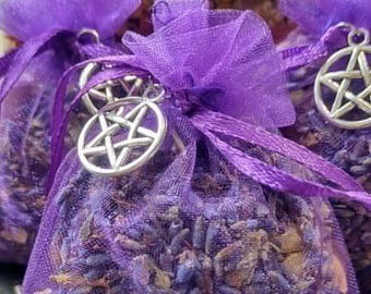 Op de afbeelding: Drie kleine, paarse gaas zakjes gevuld met gedroogde lavendel knoppen. Elke zak is vastgebonden met een paars lint en versierd met een zilveren pentagram bedel. Op de achtergrond een pot gevuld met gedroogde bloemen.