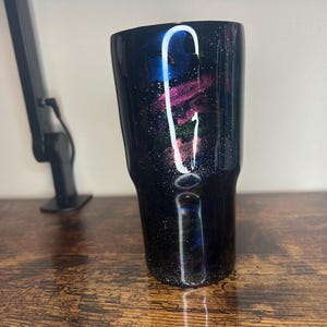 May include: A black tumbler with a galaxy-themed design. The tumbler features a blend of dark blue, purple, and pink hues, speckled with white accents. The design resembles a nebula or a starry night sky. The tumbler is made of stainless steel.