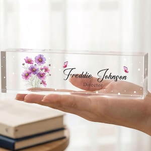 Puede incluir: Una placa de escritorio de acrílico transparente con un diseño floral de flores y mariposas moradas. El nombre "Freddie Johnson" y el título "Director" se muestran en escritura cursiva. La placa rectangular se sostiene en una mano.