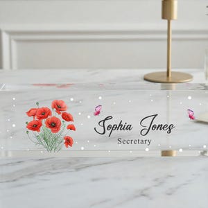 Puede incluir: Placa de escritorio de acrílico transparente con un diseño floral de amapolas rojas y hojas verdes. La placa muestra el nombre "Sophia Jones" y el título "Secretary" en escritura cursiva, con acentos de mariposa.
