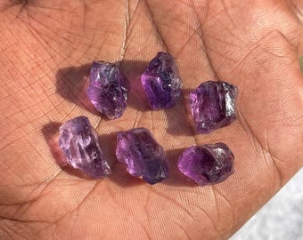 Peut inclure: Plusieurs cristaux d'améthyste bruts, d'un violet profond et d'une texture naturelle et rugueuse. Les cristaux sont présentés dans une main, révélant leurs formes et tailles variées, idéales pour la joaillerie ou la lithothérapie.