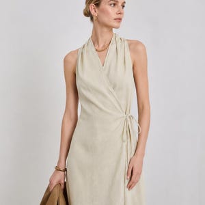 Könnte beinhalten: Beiges Wickelkleid mit Neckholder-Ausschnitt. Das Kleid ist ärmellos und hat einen Bindverschluss in der Taille. Eine braune Tragetasche wird in der linken Hand gehalten. Das Kleid ist aus leichtem, fließendem Stoff.