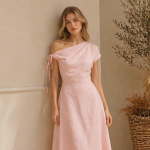 May include: A pale pink, one-shoulder midi dress with a fitted waist and a flowing skirt. The dress has a tie detail on the shoulder and is made of a soft, textured fabric. The model is wearing sandals.