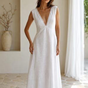 May include: A floor-length white sleeveless dress featuring a deep V-neckline with delicate lace trim detailing along the edges. The garment has a fitted bodice that transitions into a flowing, full-length skirt, crafted from a textured fabric.