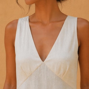 May include: A sleeveless, cream-colored dress with a deep V-neckline. The dress features a tailored bodice with seams that create a flattering shape. The fabric appears to be a lightweight linen or similar material, suitable for warm weather.
