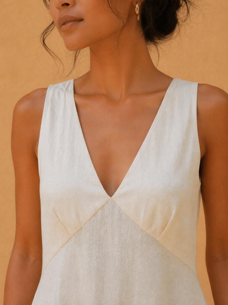May include: A sleeveless, cream-colored dress with a deep V-neckline. The dress features a tailored bodice with seams that create a flattering shape. The fabric appears to be a lightweight linen or similar material, suitable for warm weather.