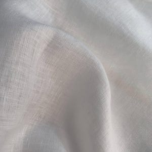 Puede incluir: Una pieza de tela blanca con una textura tejida visible y apariencia de fibra natural. El material est&aacute; drapeado para crear pliegues y sombras suaves y fluidos, resaltando la calidad semitransparente y el tejido consistente de la superficie.