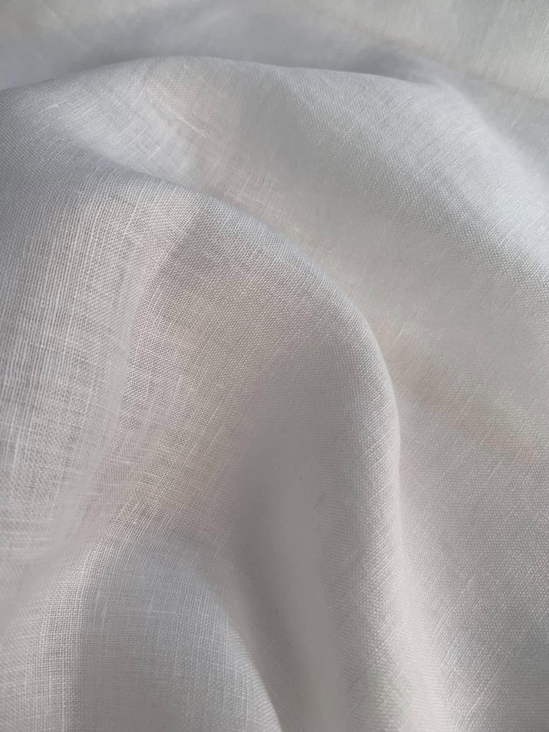 Puede incluir: Una pieza de tela blanca con una textura tejida visible y apariencia de fibra natural. El material est&aacute; drapeado para crear pliegues y sombras suaves y fluidos, resaltando la calidad semitransparente y el tejido consistente de la superficie.