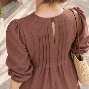 May include: A brown long-sleeved dress featuring a round neckline with a single button closure at the back, a vertical keyhole opening, and multiple pleats extending from the neckline down the back, paired with a woven straw bag with brown straps.