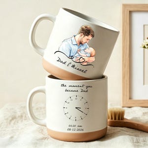 Può includere: Due tazze impilate in ceramica bianca con basi beige. La tazza superiore presenta un'illustrazione di un padre che tiene in braccio un bambino, con la scritta "Dad & Misael". La tazza inferiore ha un design a orologio e la scritta "the moment you became Dad."