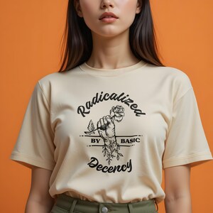 Op de afbeelding: Crèmekleurig T-shirt met de tekst "Radicalized by Basic Decency" in zwarte letters. De afbeelding toont een vuist die een roos vasthoudt, met bloemmotieven. Het shirt heeft korte mouwen en een ronde hals.