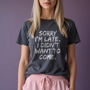 Puede incluir: Camiseta gris oscuro con el texto blanco "SORRY I'M LATE, I DIDN'T WANT TO COME". La camiseta se combina con pantalones rosas. La modelo tiene el pelo rubio y está frente a un fondo morado.