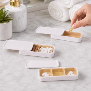 Puede incluir: Tres pastilleros rectangulares con tapas deslizantes blancas e interiores de color tostado claro. Cada recipiente cuenta con múltiples compartimentos que contienen varias pastillas blancas, cápsulas marrones y cápsulas de gel amarillas, con una tapa parcialmente abierta.