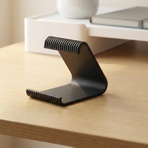 Può includere: Un supporto per telefono nero curvo con scanalature sulla parte superiore e inferiore. Il supporto è realizzato in un materiale solido ed è progettato per contenere un telefono o un tablet. Si trova su una scrivania in legno chiaro.