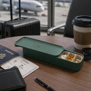 pill organiser open on an airport table with passport, boarding pass, coffee, and suitcase, plane visible through window.