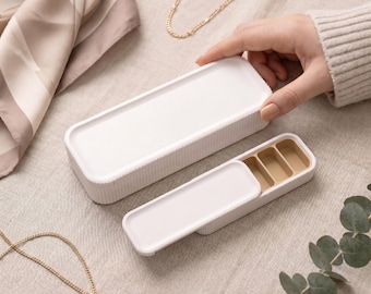 Peut inclure: Deux boîtes de rangement rectangulaires blanches avec des textures latérales nervurées. Une boîte est fermée par un couvercle solide, tandis que l'autre est entrouverte, révélant trois compartiments intérieurs beiges et un couvercle coulissant.