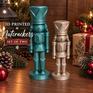 May include: A set of two 3D printed nutcracker figurines, one in metallic teal and one in metallic gold. Each nutcracker features a tall, geometric hat, a uniform-style jacket with textured details, and a circular base. The text 3D PRINTED NUTCRACKERS SET OF TWO is visible.