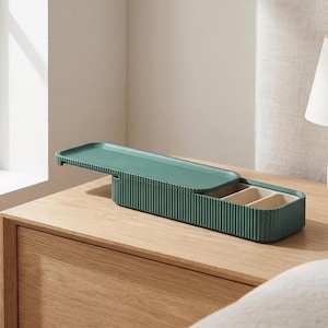 Green ribbed pill holder with sliding lid, placed open on a wooden bedside table next to a lamp.