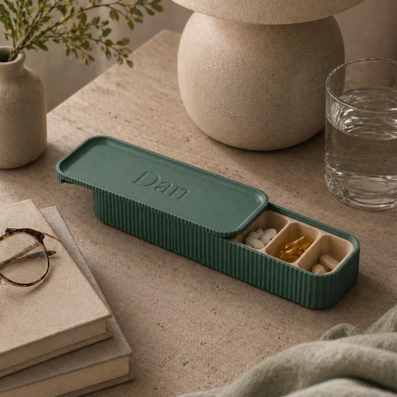 personalised pill organiser partially open on a neutral bedside table, with capsules inside, next to a glass of water, books, and soft decor.