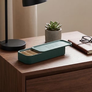 Green pill holder with “Dan” on the lid, placed on a wooden bedside table beside a lamp, plant, and notebook.