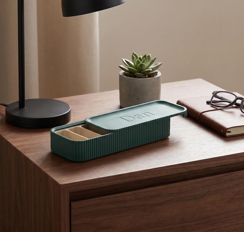 Green pill holder with “Dan” on the lid, placed on a wooden bedside table beside a lamp, plant, and notebook.