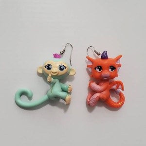 May include: A pair of dangle earrings featuring a light blue monkey and an orange dragon. Both figures have large eyes and are wearing a crown.