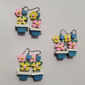Könnte beinhalten: Ein Paar Ohrringe mit einem Blumentopfdesign mit drei Blumen in jedem Topf. Die Blumen sind rosa, gelb und blau. Die Töpfe sind blau mit weißem Rand.