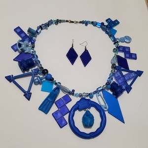 Peut inclure: Un collier bleu avec divers breloques en plastique bleu, dont un triangle, un carré, un poisson, une sucette et un anneau. Le collier comprend également des perles bleues et argentées. Le collier est accompagné d'une paire de boucles d'oreilles bleues en forme de diamant.