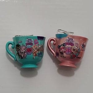 Op de afbeelding: Twee decoratieve theekopjes, één turquoise en één roze, elk met een oor en een voet. Op het turquoise kopje staat de tekst "Itty Bitty Prettys" en "over 20 surprises inside!". Op het roze kopje staat de tekst "Itty Bitty Prettys" en "over 25 surprises inside!"