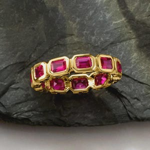 Peut inclure: Une bague en or ornée de pierres précieuses roses rectangulaires et vives, disposées en motif répétitif. La bague est présentée sur une surface sombre et texturée, mettant en valeur le design et la couleur du bijou.