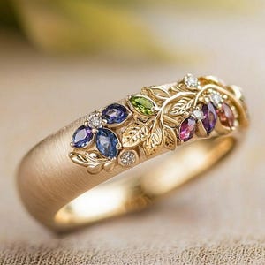 May include: A gold ring with a brushed matte finish featuring a decorative band adorned with marquise-cut gemstones in blue, green, and purple hues, interspersed with small round clear stones and sculpted gold leaf and vine metalwork details.