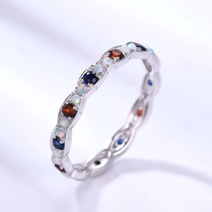 Puede incluir: Un anillo de plata con un patrón repetitivo de piedras preciosas de colores. El anillo presenta una variedad de piedras, incluyendo blancas, azules y rojas, engastadas en una banda continua. Las piedras preciosas son redondas y espaciadas.