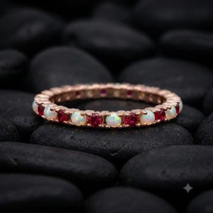 May include: A rose gold eternity band featuring an alternating pattern of round, iridescent white opals and vibrant, deep red rubies set in a continuous circle. The metal band has a polished finish, showcasing the contrast between the gemstones.