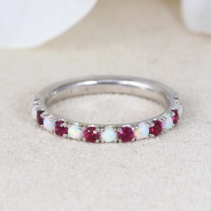 May include: A silver band ring featuring alternating white opal and red ruby gemstones. The ring is set on a neutral background, showcasing the vibrant colors of the gemstones. The ring is a delicate piece of jewelry.