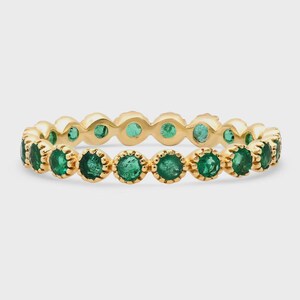 May include: A gold ring featuring a continuous row of round, vibrant green emeralds. The ring's band is a polished gold, with each emerald set in a delicate, scalloped bezel. The gemstones are evenly spaced around the entire band.
