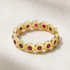 May include: A gold ring featuring a series of small, round, ruby-colored gemstones set in individual bezels. The ring has a smooth, polished finish and a classic design, suitable for everyday wear or special occasions.