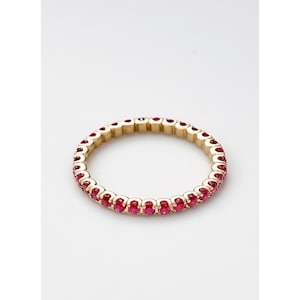 Peut inclure: Une bague en or ornée d'une rangée continue de pierres précieuses rouges vives de forme ovale. La bague est mise en valeur sur un fond blanc, soulignant le contraste entre l'or et les pierres rouges. Le design de la bague est simple et élégant.