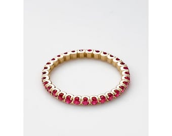 May include: A gold ring with a continuous row of oval-cut, vibrant red gemstones. The ring is set against a white background, highlighting the contrast between the gold and the red stones. The ring's design is simple and elegant.