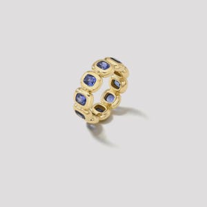 May include: A gold ring featuring a series of square-cut, blue gemstones. The ring has a continuous design with each gemstone set in a gold bezel. The ring is displayed on a white background, showcasing its polished finish and intricate details.