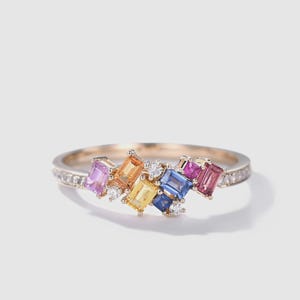 Pode incluir: Um anel de ouro com um aglomerado de pedras preciosas retangulares coloridas em tons de rosa, laranja, amarelo, azul e roxo. Pequenas pedras brancas são intercaladas entre as gemas maiores, e a banda também é adornada com pequenas pedras brancas.
