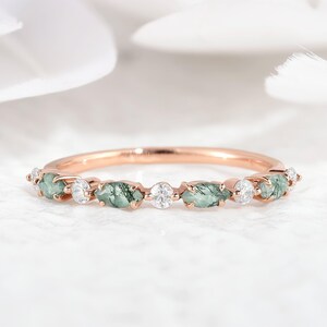 May include: A rose gold ring featuring a series of green moss agate stones and small, round, clear gemstones. The ring's band is thin and polished, with the gemstones set in a row along the top.