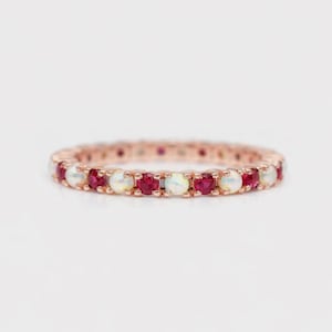 May include: A rose gold eternity ring with alternating red gemstones and white opals. The ring has a continuous row of round-cut stones. The ring is displayed on a white background. The gemstones and opals create a full circle of colour and sparkle.